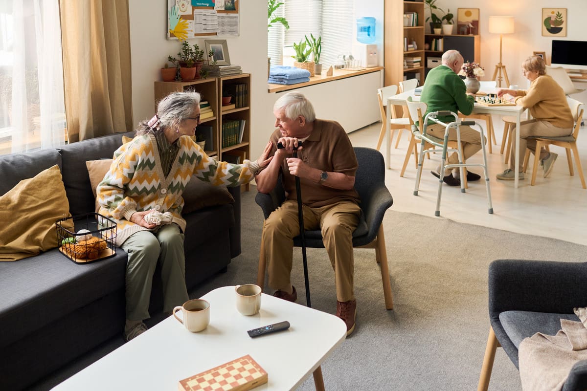 Senior residents socializing in a care home, one couple talking on a sofa while another plays a board game